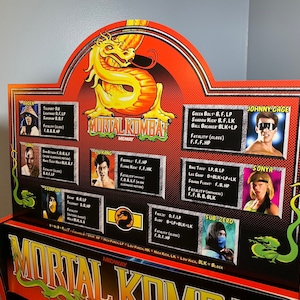 May include: A Mortal Kombat arcade game cabinet with a red and black design. The cabinet features a green dragon and the text "Mortal Kombat Midway" on the front. The cabinet also has a character selection screen with images of the characters and their special moves.