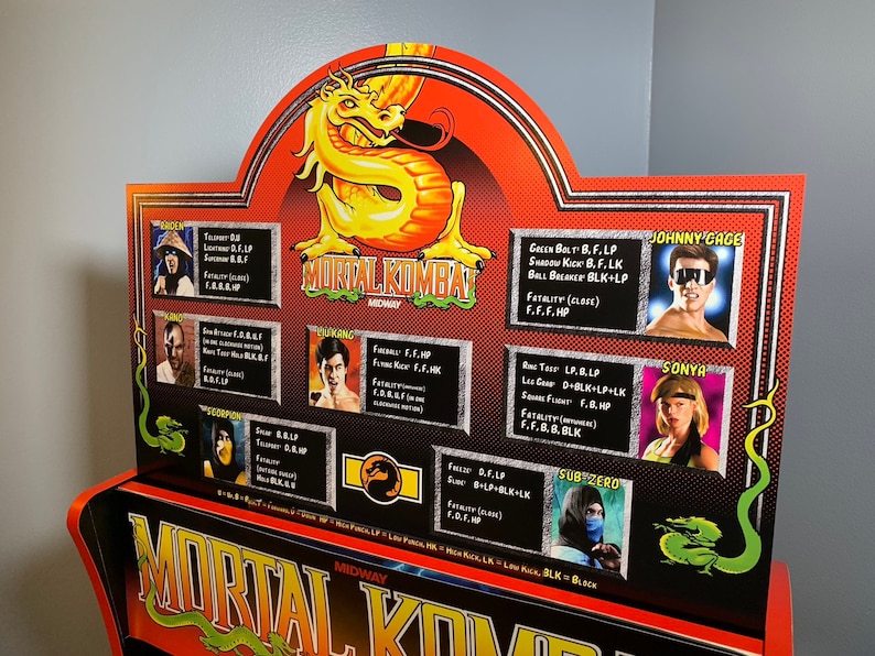 May include: A Mortal Kombat arcade game cabinet with a red and black design. The cabinet features a green dragon and the text "Mortal Kombat Midway" on the front. The cabinet also has a character selection screen with images of the characters and their special moves.