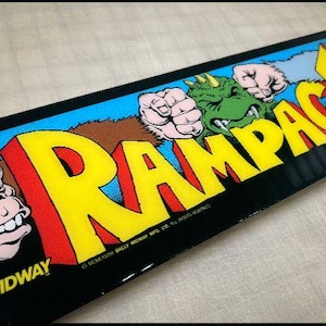 May include: A black and yellow arcade game cabinet marquee with the text "Rampage" in large, bold letters. The marquee features a green dinosaur, a gorilla, and a werewolf.