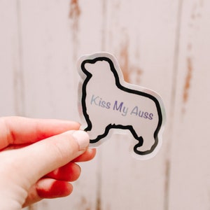 May include: A clear sticker with a black outline of an Australian Shepherd dog. The sticker has the text "Kiss My Aussies" in a purple and blue gradient.