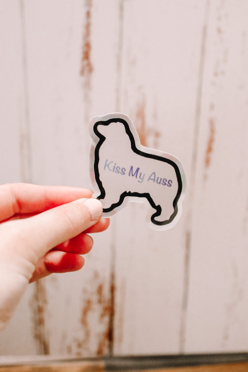 May include: A clear sticker with a black outline of an Australian Shepherd dog. The sticker has the text "Kiss My Aussies" in a purple and blue gradient.