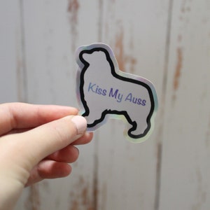 May include: A holographic sticker with a black outline of an Australian Shepherd dog. The sticker says "Kiss My Aussies" in purple text.