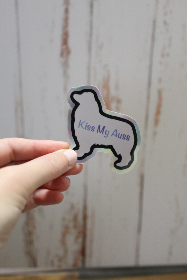 May include: A holographic sticker with a black outline of an Australian Shepherd dog. The sticker says "Kiss My Aussies" in purple text.