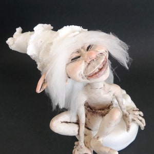 May include: A white ceramic figurine of a smiling gnome with a white fluffy hat and long white hair. The gnome is sitting on a white base.