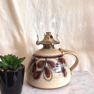May include: A ceramic oil lamp with a rounded base featuring a cream-colored glaze and brown leaf-shaped patterns. The lamp has a brass burner assembly and a clear glass chimney. Beside it sits a small succulent in a glossy black pot.