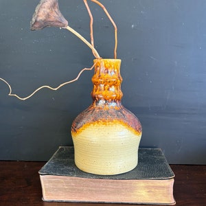 May include: A brown and beige ceramic vase with a textured surface. The vase is sitting on top of a black and brown book.