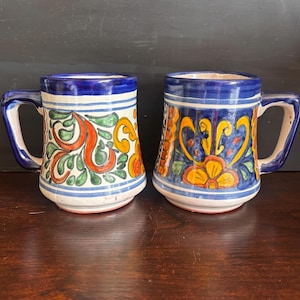 May include: Two ceramic mugs with blue handles and rims. The mugs feature hand-painted designs in orange, yellow, and green on a white background. The mugs have a traditional, rustic style, perfect for coffee or tea.