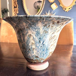 May include: A handcrafted ceramic vase with a flared rim. The vase features a blend of blue, white, and brown hues, creating a marbled effect. It sits on a small, round, beige base. The vase is a decorative item.
