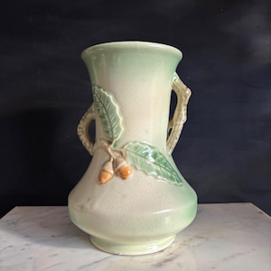 May include: A light green and cream-colored ceramic vase with a detailed acorn and leaf design. The vase has two branch-like handles and is approximately 10 inches tall. The design features green leaves and orange acorns.