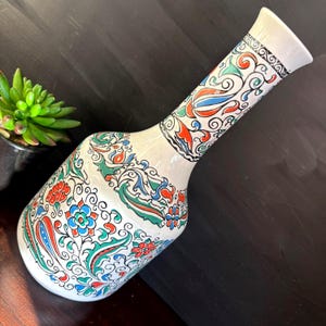 May include: A white ceramic vase with a long neck and a bulbous body. The vase is adorned with a colorful floral pattern in red, blue, green, and orange. The rim of the vase has a black and white geometric design.
