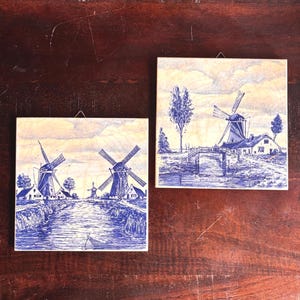 May include: Two square decorative tiles featuring blue and white illustrations of Dutch windmills and rural landscapes. The artwork depicts windmills, houses, water, and trees under a cloudy sky, evoking a sense of traditional Dutch scenery.