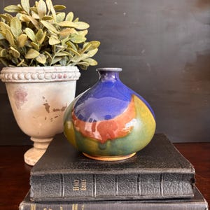 May include: A colorful ceramic vase with a rounded body and a narrow neck. The vase features a vibrant design with blue, green, orange, and yellow hues. It sits atop a stack of old books, with a potted plant in the background.