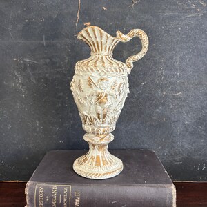 May include: A white ceramic pitcher with gold accents and an embossed cherub design. The pitcher has a wide mouth and a handle that curves up and over the top.