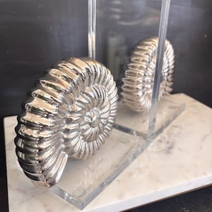 May include: Silver-colored, nautilus shell bookends. The bookends are made of a shiny, metallic material and are set in clear acrylic holders. The base is a white marble with gray veining. The bookends are designed to hold books upright.