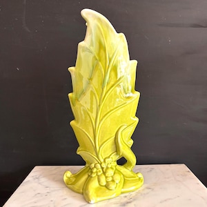 May include: A tall, lime green ceramic vase featuring a stylized leaf design with raised veins and a cluster of small, rounded berry-like shapes at the base. The glossy finish reflects light, highlighting the vertical, pointed structure of the piece.