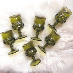 May include: Six olive green vintage goblets with a textured pattern on the base and a smooth, rounded bowl. The goblets are arranged on a white, fluffy surface, showcasing their unique design and color.