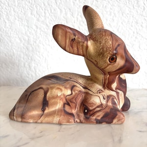 May include: A ceramic deer figurine in shades of brown and tan, with a marbled effect. The deer is in a resting position, with its head and ears upright. The figurine has a glossy finish and is set against a white background.