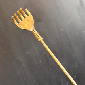 May include: A gold-toned metal back scratcher featuring a hand-shaped head with five distinct, rounded tines. The head is attached to a long, slender, cylindrical handle that extends diagonally across the frame, showcasing a smooth, metallic finish.