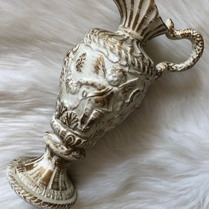 May include: White ceramic pitcher with gold accents and intricate detailing. The pitcher has a wide mouth and a handle shaped like a snake.