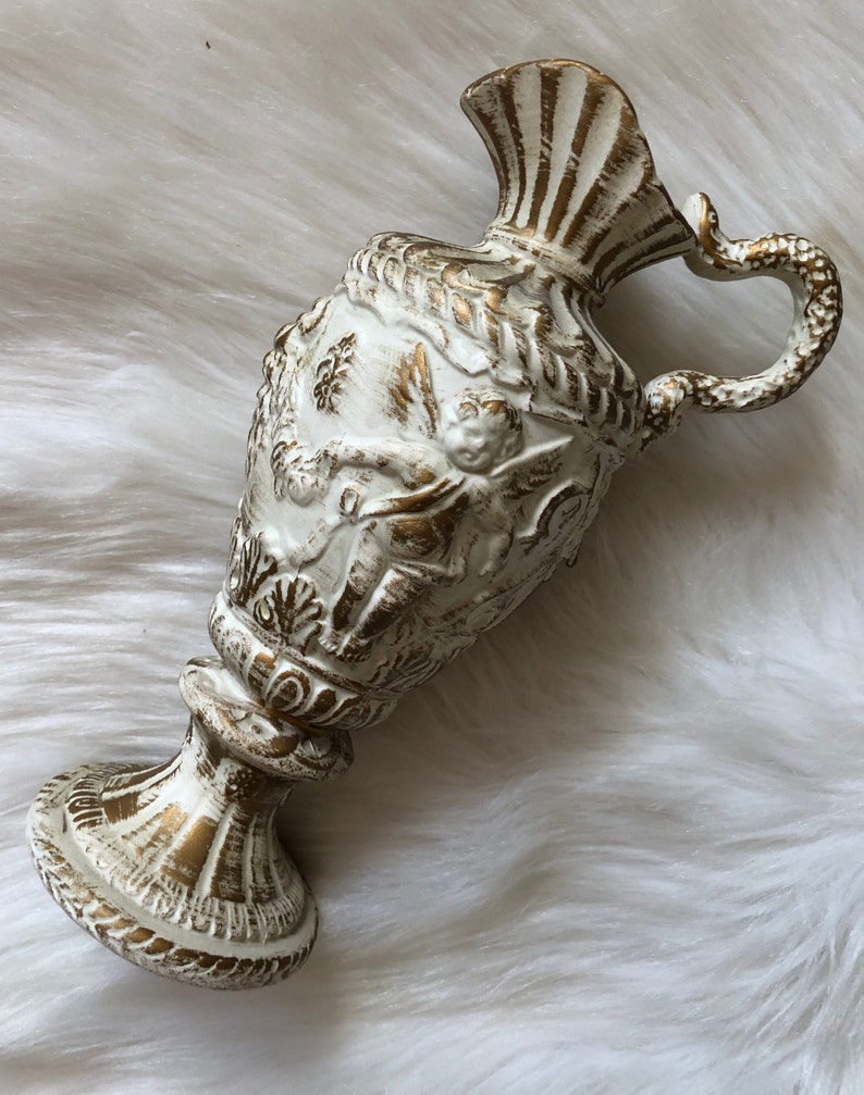 May include: White ceramic pitcher with gold accents and intricate detailing. The pitcher has a wide mouth and a handle shaped like a snake.