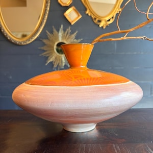 May include: A ceramic vase with a wide, rounded body featuring a pale pink, brushed finish on the lower section and a vibrant orange, glossy upper section with a subtle fan pattern. A thin, bare branch extends from the narrow, flared neck.