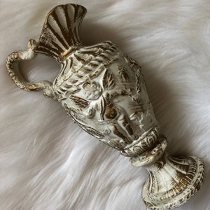 May include: White ceramic pitcher with gold accents and a cherub design. The pitcher has a wide base and a handle that curves around the top.
