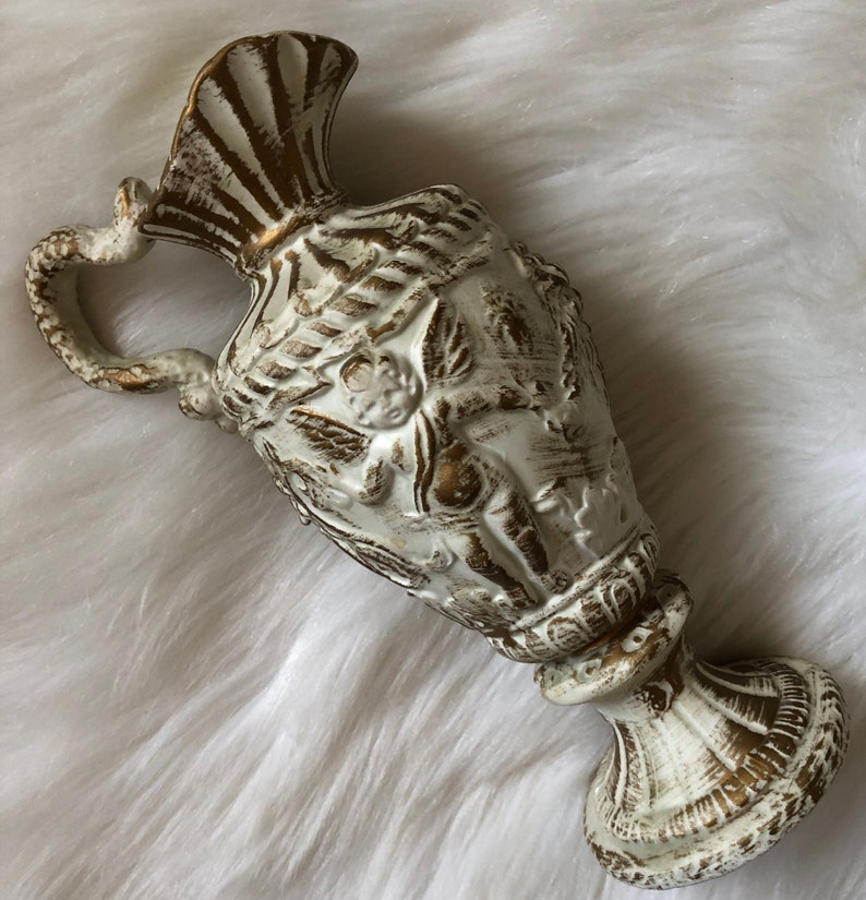 May include: White ceramic pitcher with gold accents and a cherub design. The pitcher has a wide base and a handle that curves around the top.