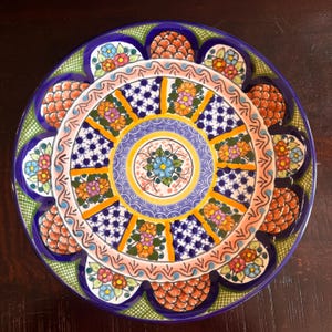 May include: A decorative ceramic plate with a vibrant floral design. The plate features a blue rim, a green border, and a central pattern with orange, yellow, pink, and blue flowers. The plate's diameter is approximately 12 inches.