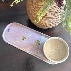 May include: A handmade ceramic serving platter and bowl set. The platter is oblong with a pink and white speckled glaze, accented with yellow and blue abstract designs. The attached bowl is a speckled beige. The set is ideal for serving appetizers or snacks.