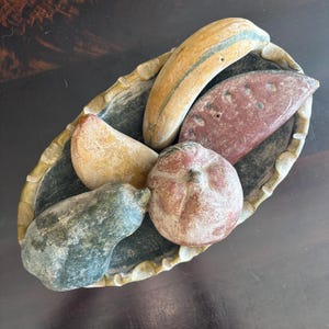 May include: A decorative oval-shaped dish filled with carved stone fruits. The fruits include a banana, watermelon slice, pear, apple, and a gourd. The dish has a scalloped edge and a dark interior.
