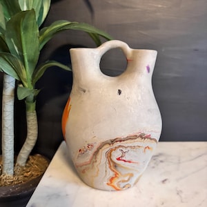May include: A cream-colored ceramic vase with a unique handle and a circular opening. The vase features colorful abstract patterns in orange, red, and brown. The vase is displayed on a white marble surface.