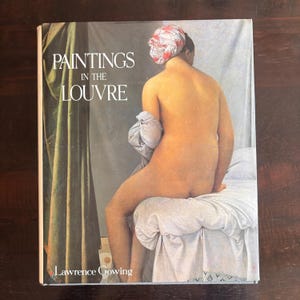 May include: A book titled "Paintings in the Louvre" by Lawrence Gowing. The cover features a nude figure seated on a bed, with a patterned head covering and a draped cloth. The background includes a green curtain.
