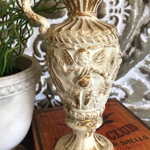 May include: A white ceramic pitcher with gold accents and a cherub design. The pitcher has a handle shaped like a snake and a base with a decorative design.