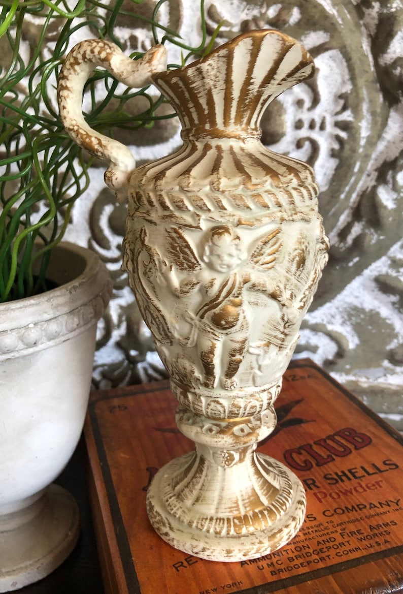 May include: A white ceramic pitcher with gold accents and a cherub design. The pitcher has a handle shaped like a snake and a base with a decorative design.