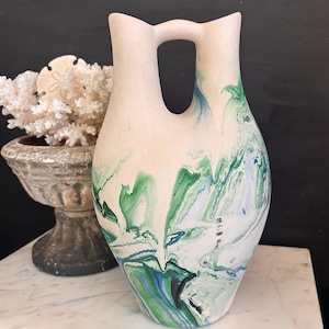 May include: A tall, off-white ceramic vase with a unique handle and a marbled design. The vase features flowing patterns of green, blue, and black against a white background. The vase is displayed on a marble surface.