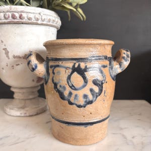 May include: A tan stoneware crock with two handles and a dark blue floral design. The antique jar has a cylindrical shape with a slightly flared rim and is decorated with horizontal blue bands.
