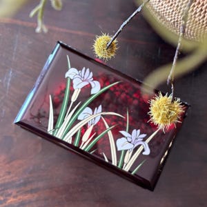 May include: A rectangular, burgundy-colored lacquer box with a floral design. The box features white irises with green stems and leaves. The background has a red, textured pattern. The box is on a dark wooden surface.