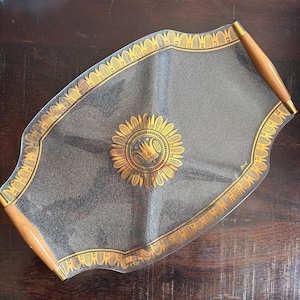 May include: A rectangular glass serving tray with wavy edges, featuring a textured surface and gold-colored decorative borders. The center displays a gold sunburst motif with a crown icon. Two smooth, light-brown wooden handles are attached at each end.