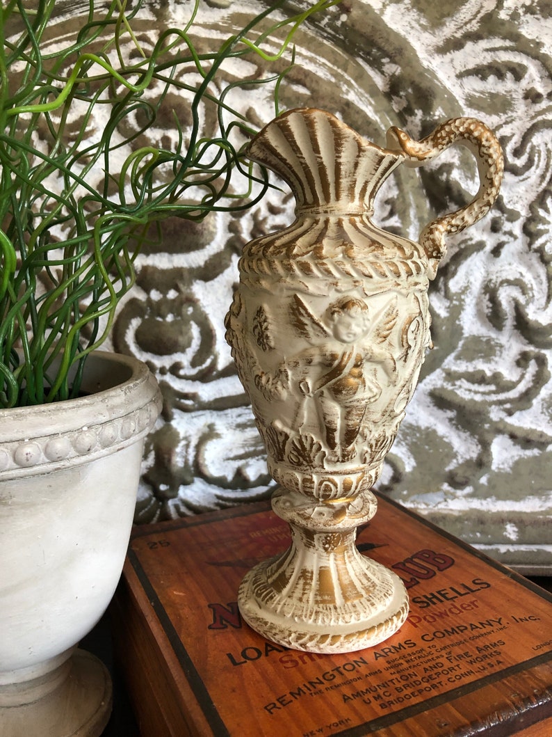 May include: A white ceramic pitcher with gold accents and a cherub design. The pitcher has a wide mouth and a handle. It is sitting on a wooden box with the text "Remington Arms Company, Inc." printed on it.