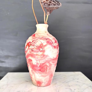 May include: A decorative vase with a swirling pattern of red and white. The vase has a narrow neck and a wider base. Dried stems and a seed pod are placed inside the vase. The vase is sitting on a white marble surface.