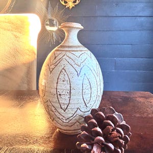 May include: A beige ceramic vase with a rounded body and a narrow neck. The vase features a repeating pattern of brown, stylized leaf shapes. A large pine cone sits in front of the vase on a wooden surface.