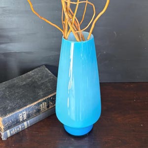 May include: A tall, light blue glass vase with a flared top and a round base. The vase contains several thin, brown branches. The vase is sitting on a dark wooden surface next to a book.