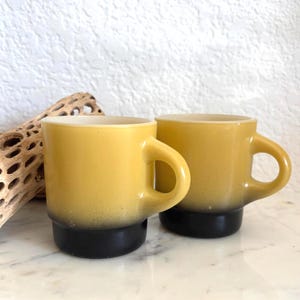 May include: Two yellow mugs with black bases are displayed. The mugs have a rounded handle and a speckled gradient effect, transitioning from yellow to black. The mugs are likely for coffee or tea.