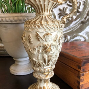 May include: A white and gold ceramic pitcher with a cherub design. The pitcher has a fluted top and a handle that curves back towards the body. The base is round and has a decorative design.
