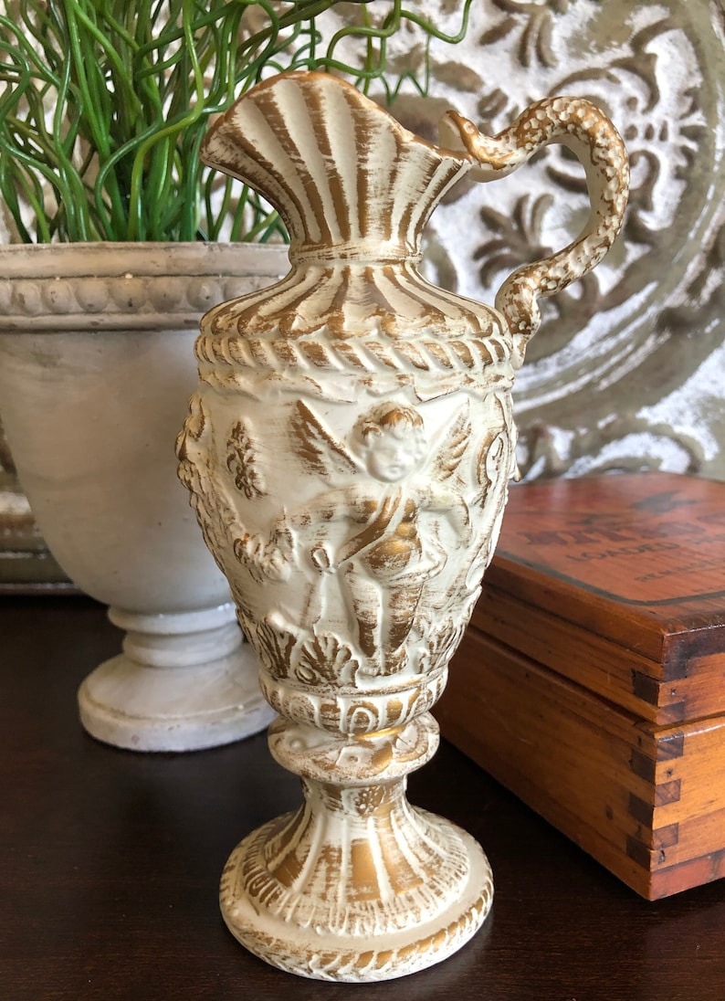 May include: A white and gold ceramic pitcher with a cherub design. The pitcher has a fluted top and a handle that curves back towards the body. The base is round and has a decorative design.