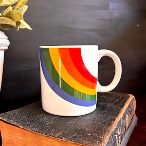 May include: White ceramic mug with a rainbow design in red, orange, yellow, green, and blue. The mug has a curved handle and is sitting on a dark book. The rainbow design is a prominent feature, adding a pop of color.