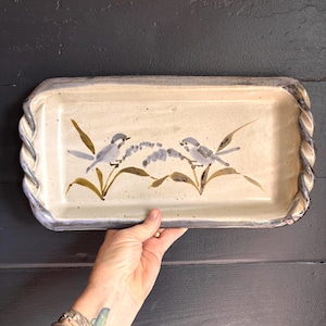 May include: A rectangular ceramic serving tray with a cream-colored base and a twisted, rope-like rim in a dark, mottled finish. The center features a hand-painted illustration of two small birds perched on leafy branches with soft blue and brown tones.