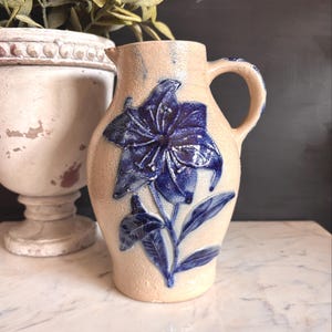May include: A beige ceramic pitcher with a blue floral design. The pitcher has a handle and a spout. The floral design is a large, stylized flower with detailed petals and leaves. The pitcher is likely vintage or antique.