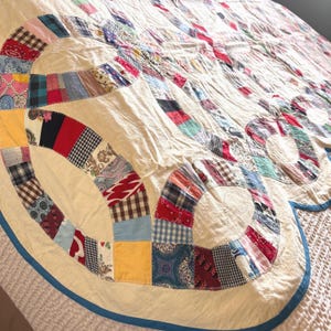 May include: A patchwork quilt featuring a double wedding ring pattern with interlocking circles made of various multicolored fabric scraps. The quilt has a cream-colored background and is finished with a solid blue fabric border trim.