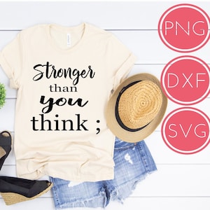 Pode incluir: Uma camiseta bege com o texto "Stronger than you think;" impresso em preto. A camiseta está colocada sobre uma superfície de madeira branca com um par de sandálias pretas, um chapéu de palha e calções de ganga.